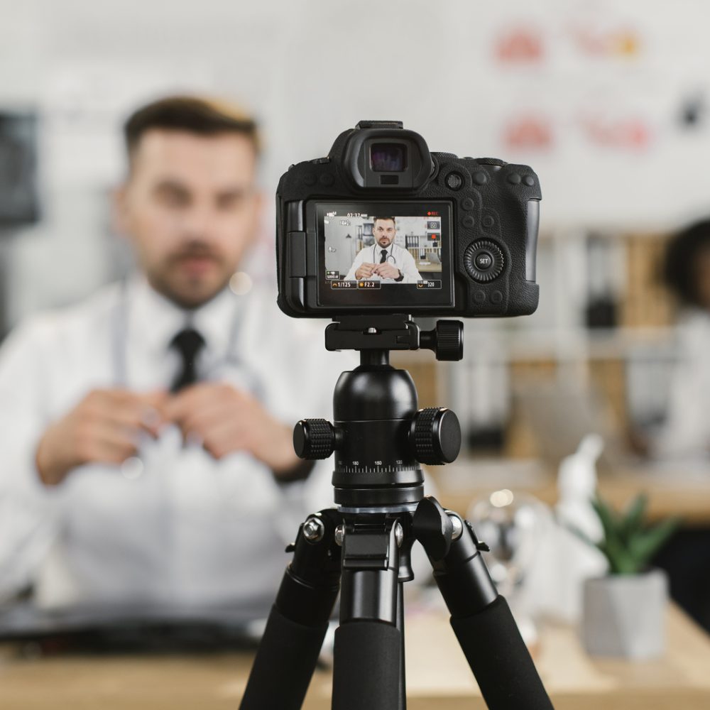 Competent medical worker in uniform doing live streaming while sitting at cabinet. Focus on screen of digital camera. Broadcast on medical theme.