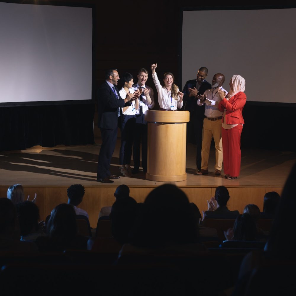 businesswoman-standing-at-the-stage-of-the-auditorium-with-colleagues-in-front-of-audience.jpg